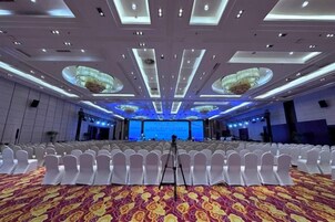 Un salón de conferencias amplio con filas de sillas blancas, un escenario con fondo azul y una alfombra estampada.
