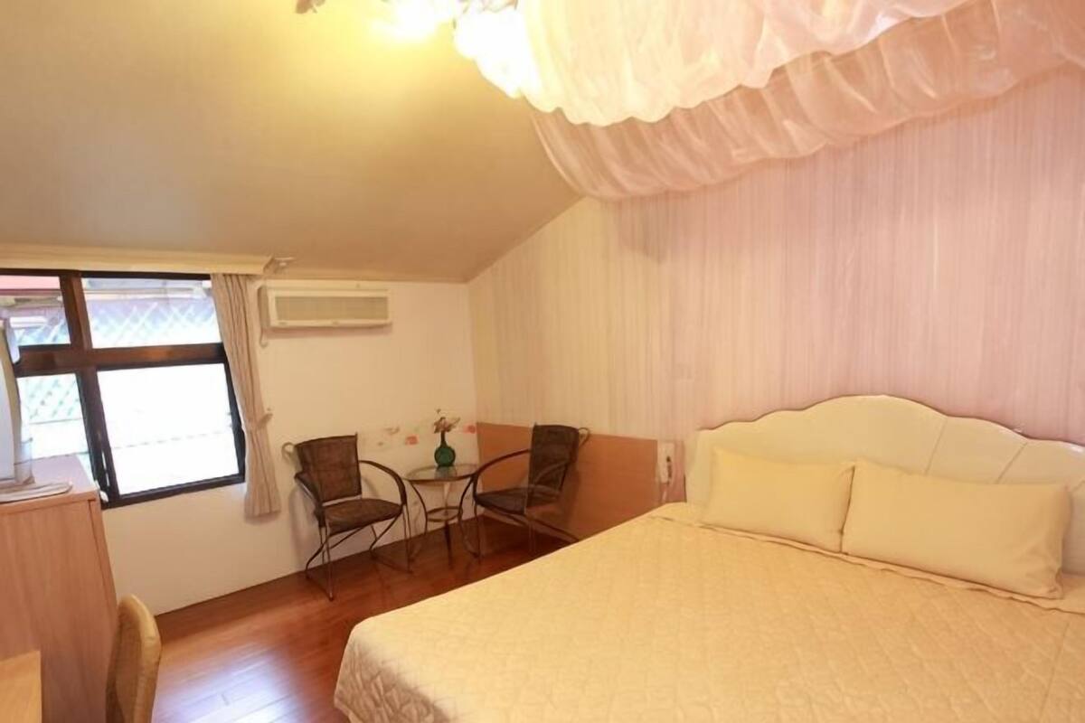 Ein Schlafzimmer mit einem Bett, zwei Stühlen, einem kleinen Tisch und einem Fenster mit Vorhängen.