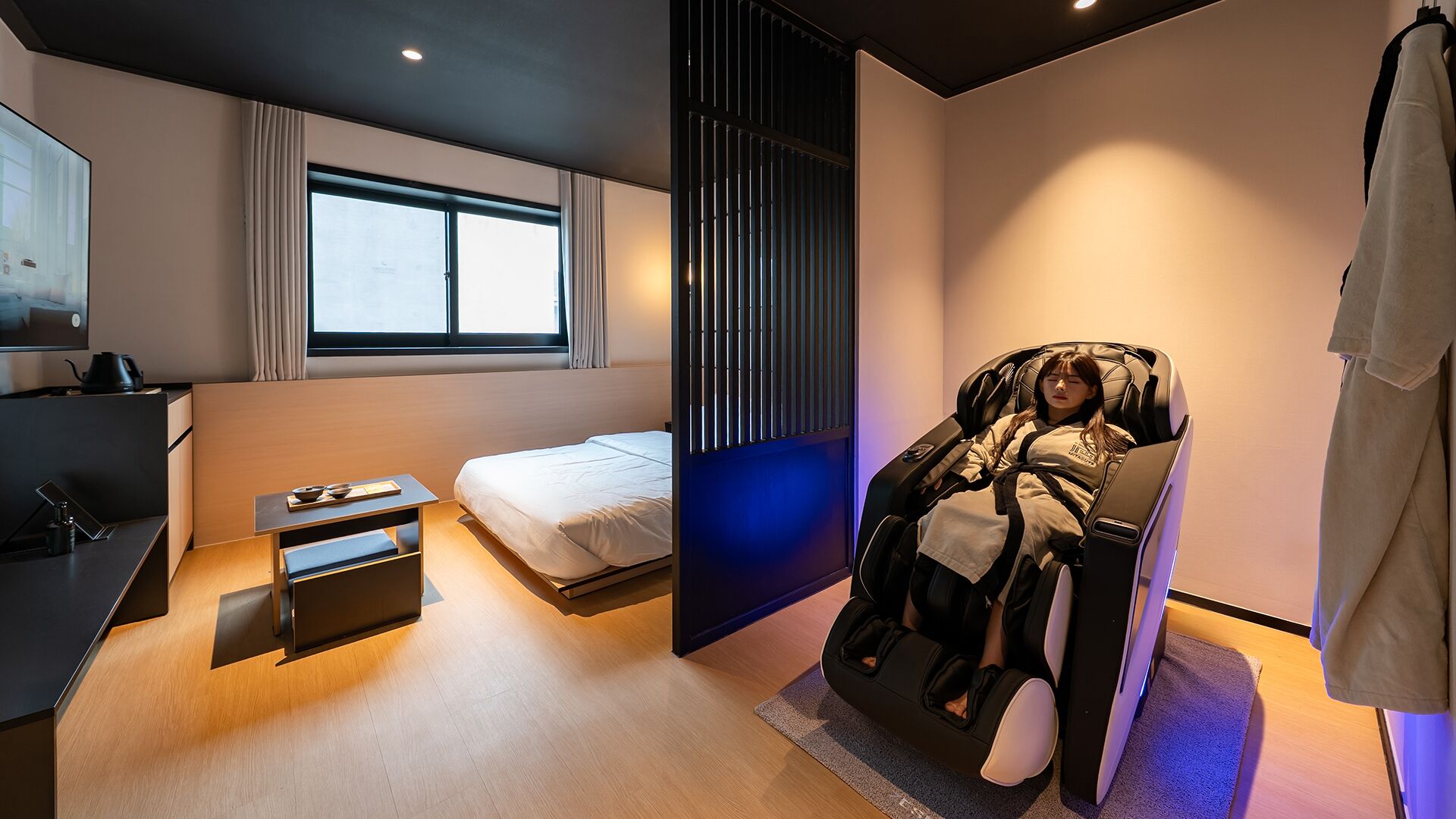 A modern hotel room with a bed, a desk, a massage chair, and a large window.