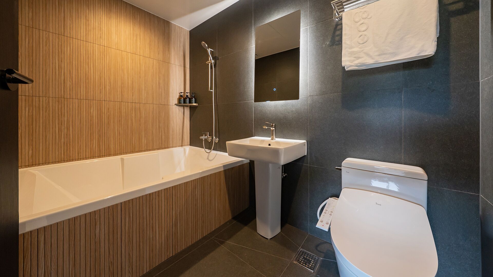 Een moderne badkamer met een houten wastafelmeubel, een witte badkuip, een zuilwastafel en een toilet.