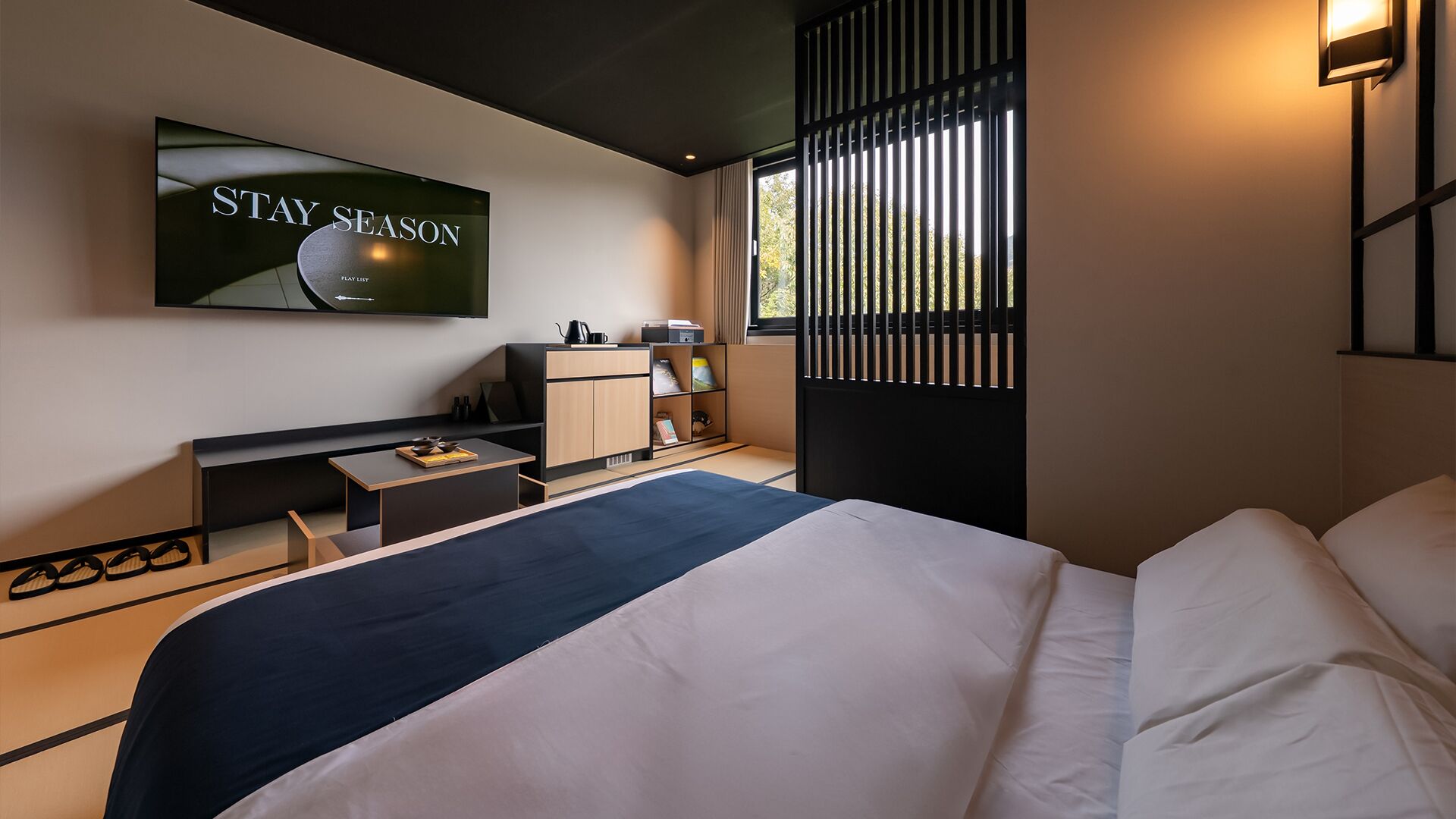 A modern hotel room with a bed, a desk, a TV displaying "STAY SEASON," and a window with blinds.