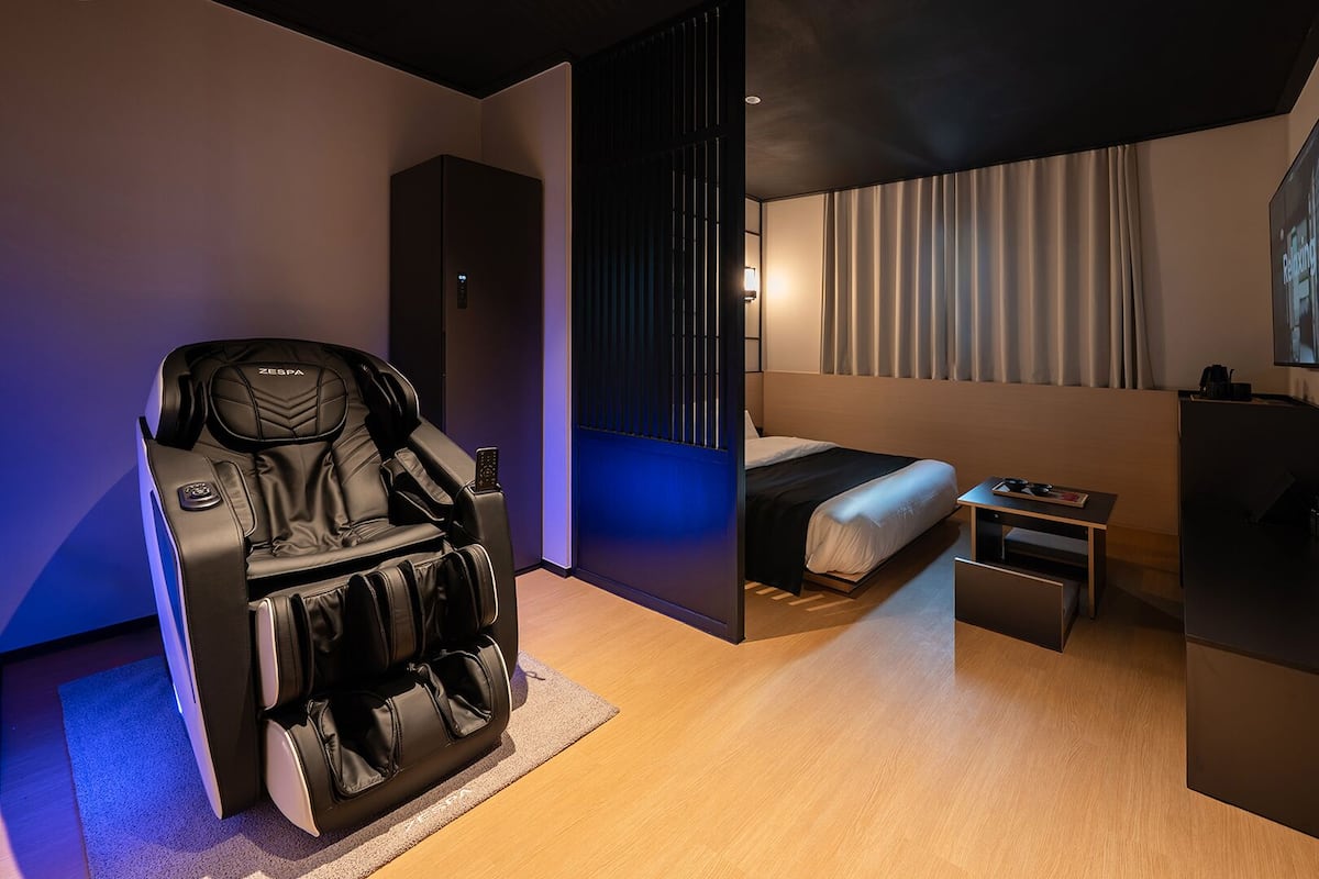 Ett modernt hotellrum med en massagestol, en säng, ett litet bord och en tv som är monterad på väggen.