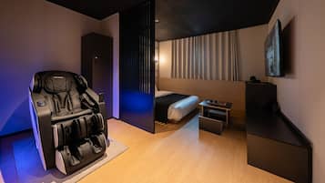 Una habitación de hotel moderna con un sillón de masaje, una cama, una mesita y un televisor montado en la pared.