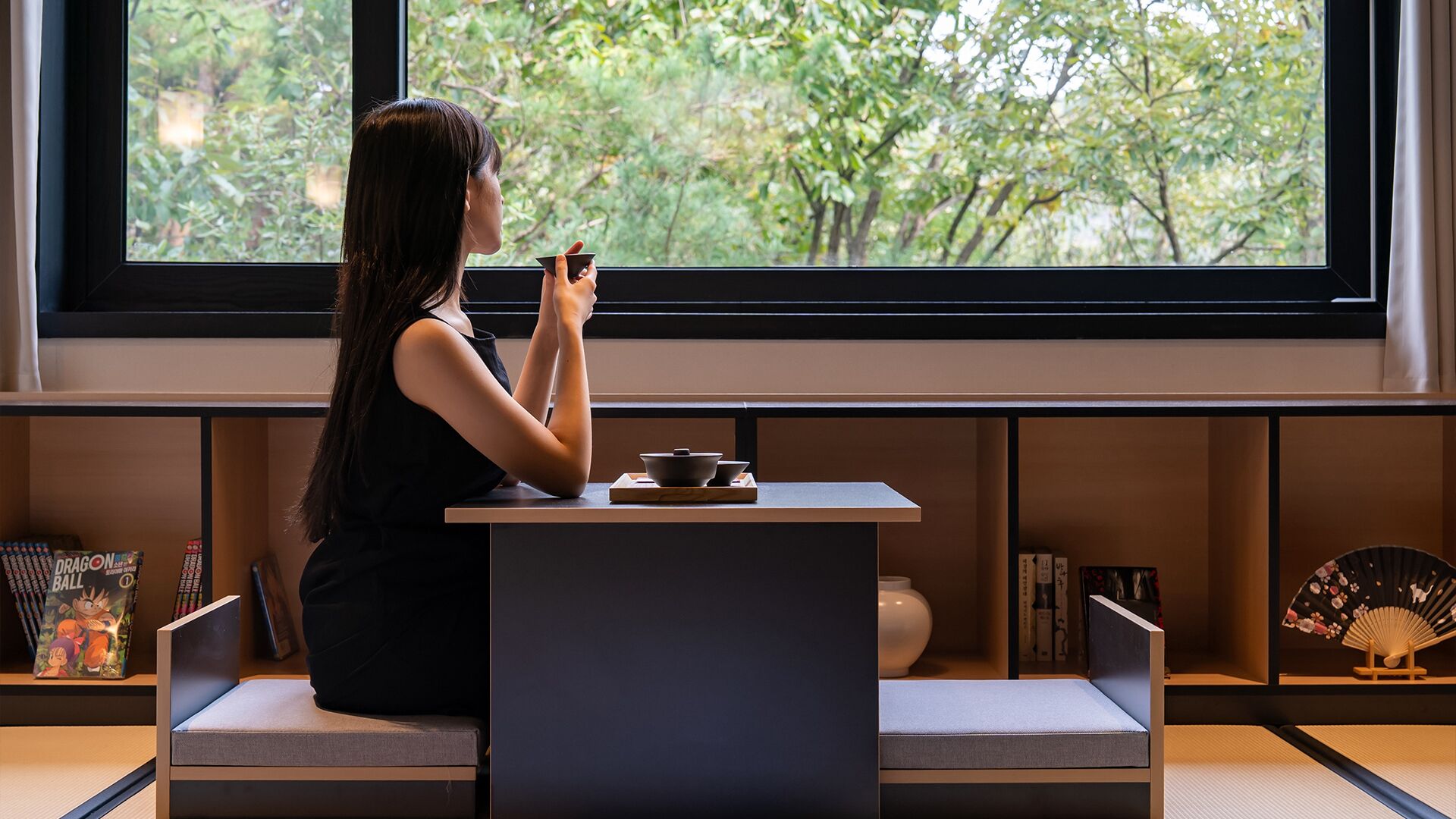 A person sitting at a table by a window with a view of trees, a fan, and a small shelf with items.
