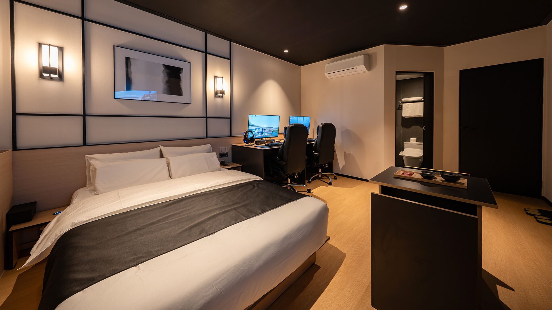 A modern hotel room with a large bed, a desk with two monitors, and a small kitchenette area.