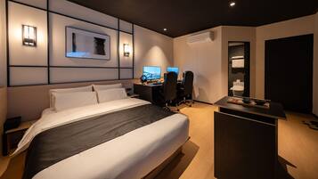 A modern hotel room with a large bed, a desk with two monitors, and a small kitchenette area.