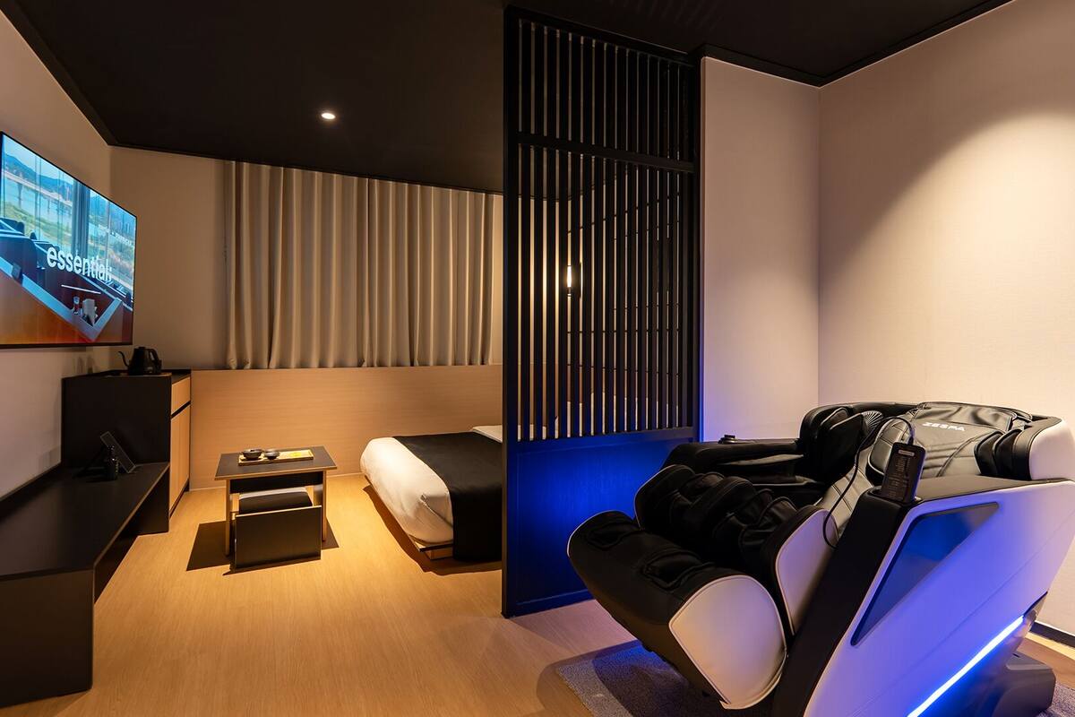 Ett modernt hotellrum med en stor platt-TV, en massagefåtölj, ett litet bord med en skål och ätpinnar samt en skjutdörr med vertikala persienner.