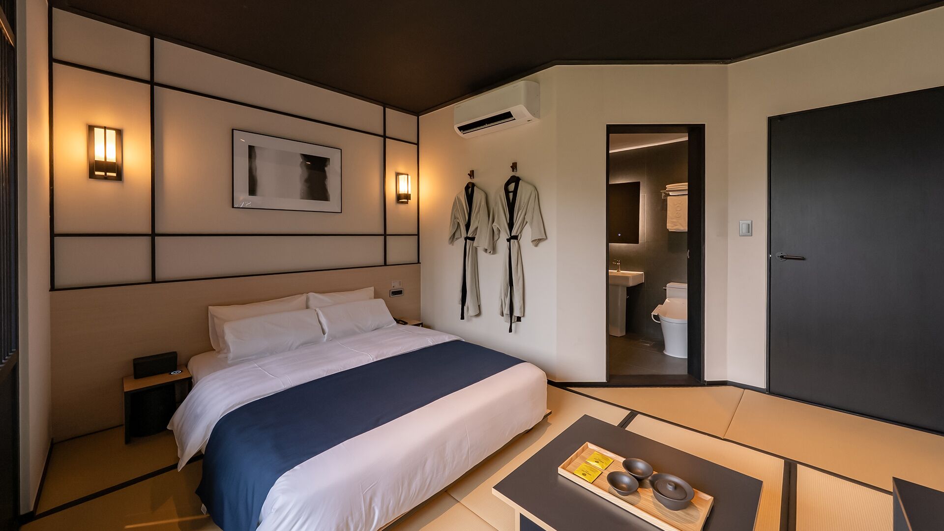 A hotel room with a bed, bedside table, wall-mounted robe hooks, a small table with tea set, and a door leading to a bathroom.