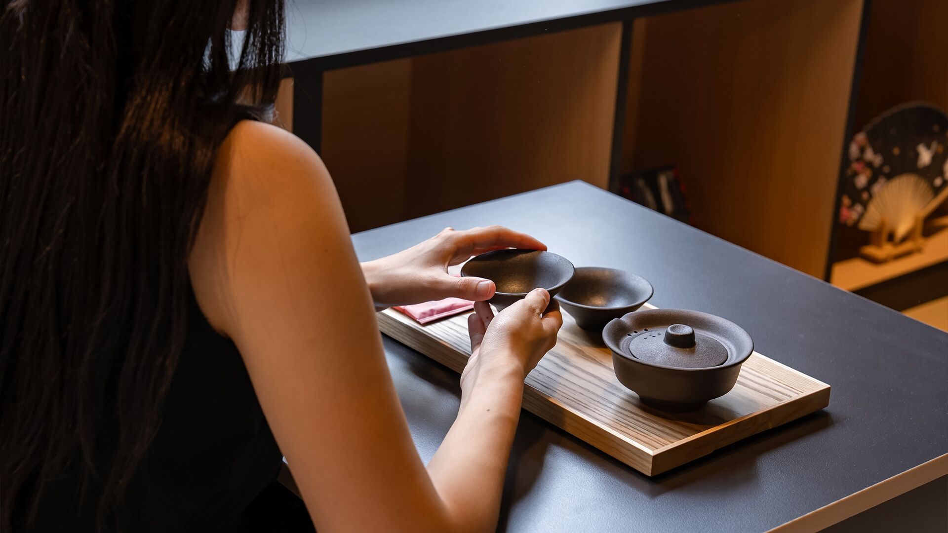A person holding a small bowl on a wooden tray with a larger bowl and a pot on a table.