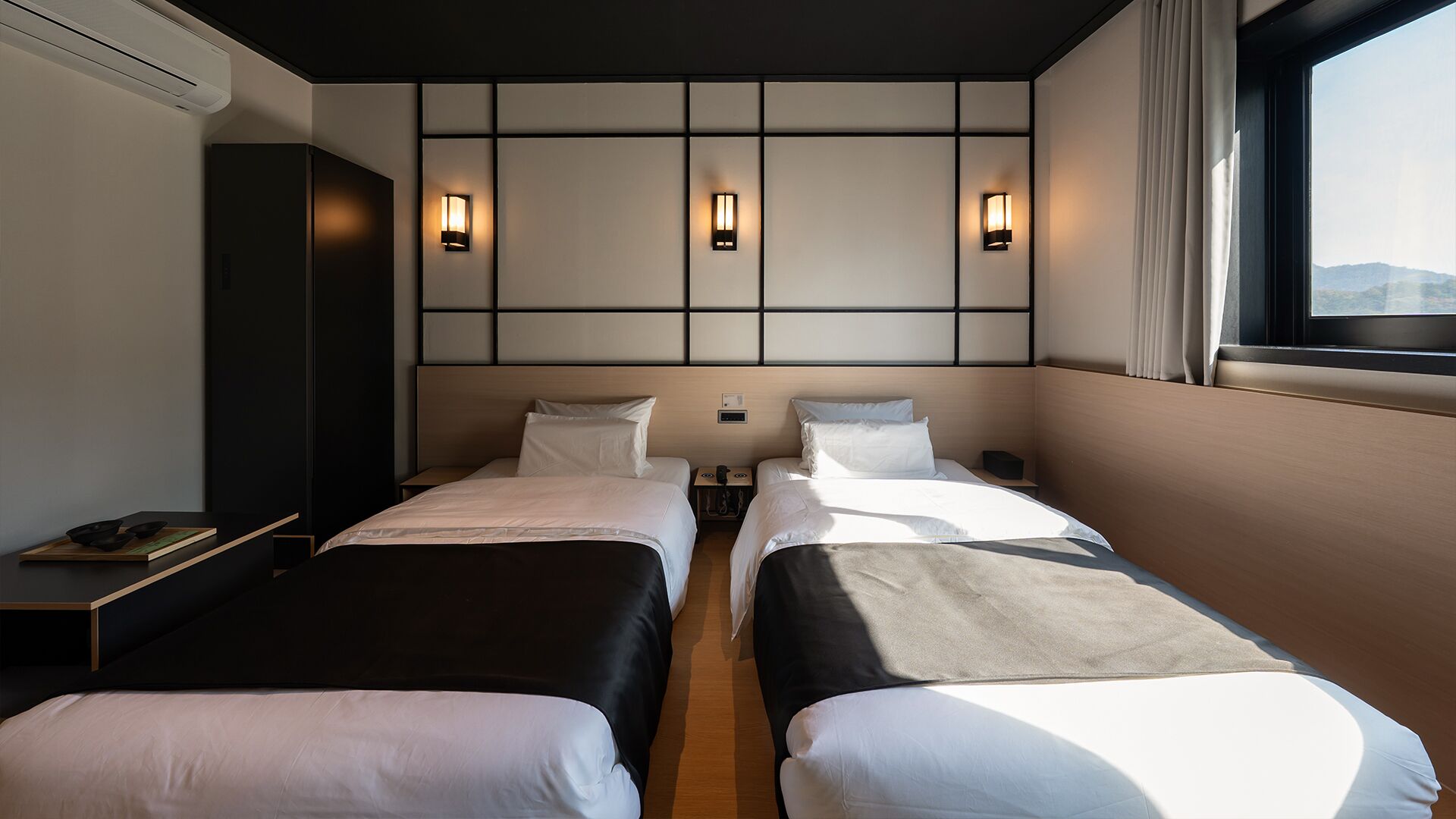 Une chambre d’hôtel avec deux lits, une table de chevet, une lampe fixée au mur et une fenêtre avec des rideaux.