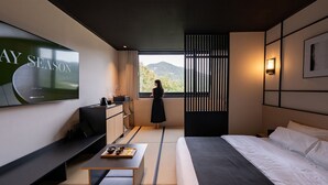 A modern hotel room with a large TV displaying 'PLAY LIST', a bed, a desk with a laptop, a small table with bowls, and a view of mountains through a window. - Mitasuya Ryokan Hotel by Anook Gongju  (Gongju)