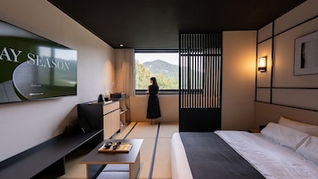 A modern hotel room with a large TV displaying 'PLAY LIST', a bed, a desk with a laptop, a small table with bowls, and a view of mountains through a window.