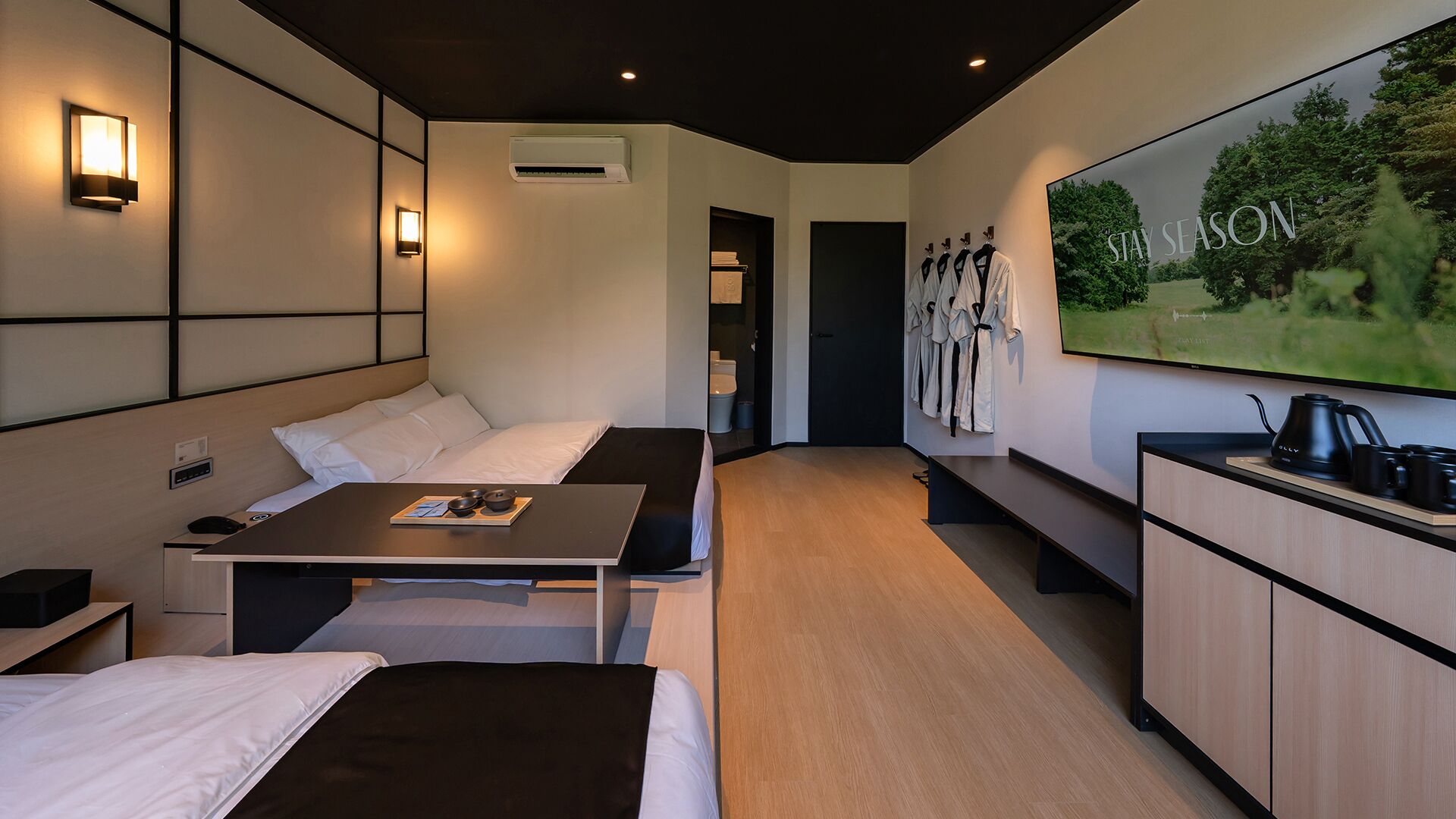 Quarto de hotel moderno com cama, mesa de cabeceira e uma grande tela de TV exibindo 'STAY SEASON'.