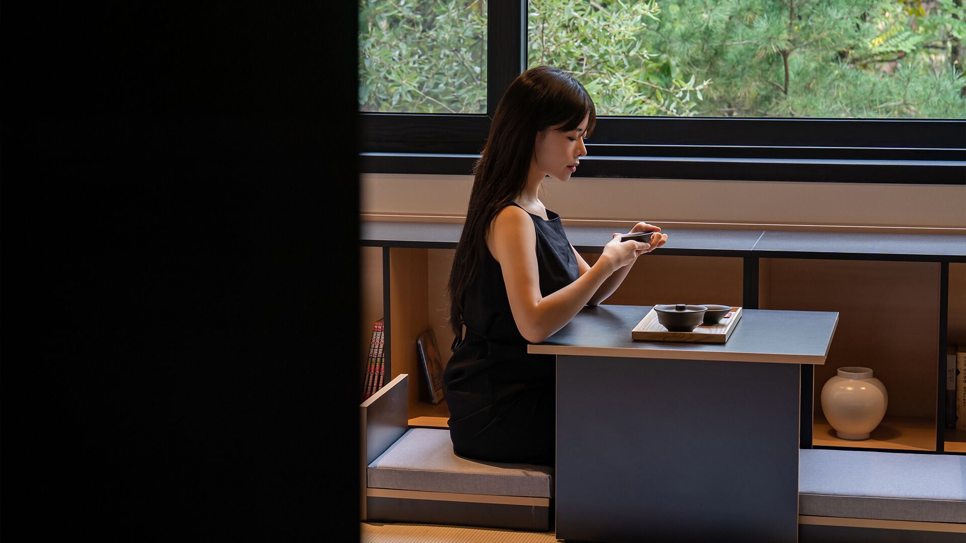 A person seated at a table with a tea set, in a room with a large window and a bookshelf.