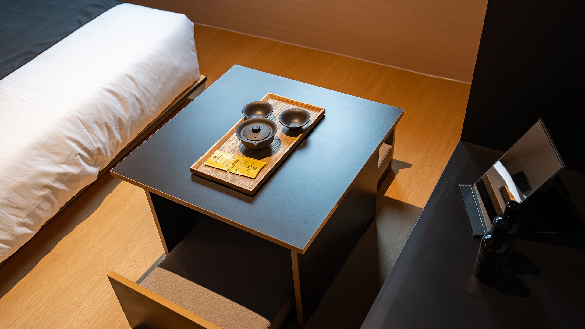 A neatly made bed with a white pillow and a wooden nightstand beside it. On the nightstand, there is a tray with a teapot, two cups, and a tea bag.