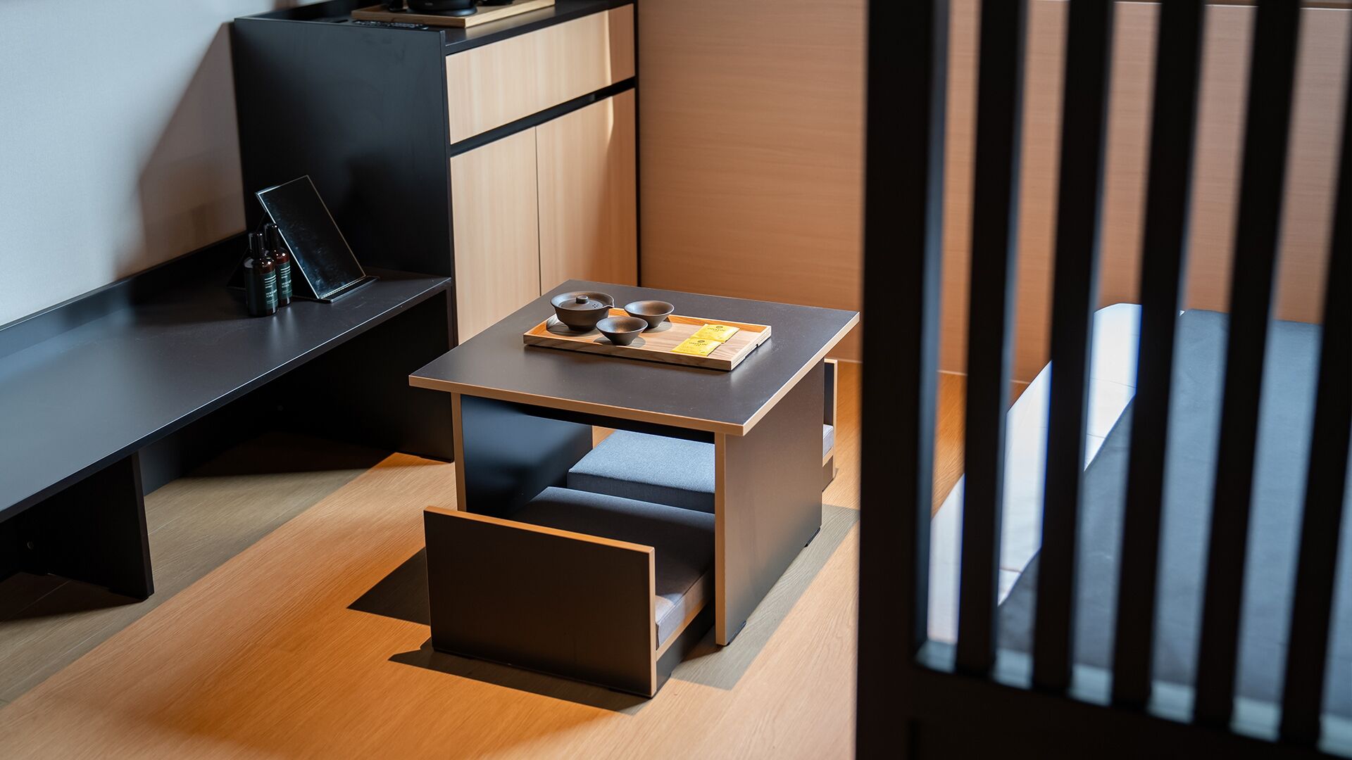 A modern, minimalist room with a wooden floor, a small table with a tray of tea utensils, a black desk, and a black cabinet.