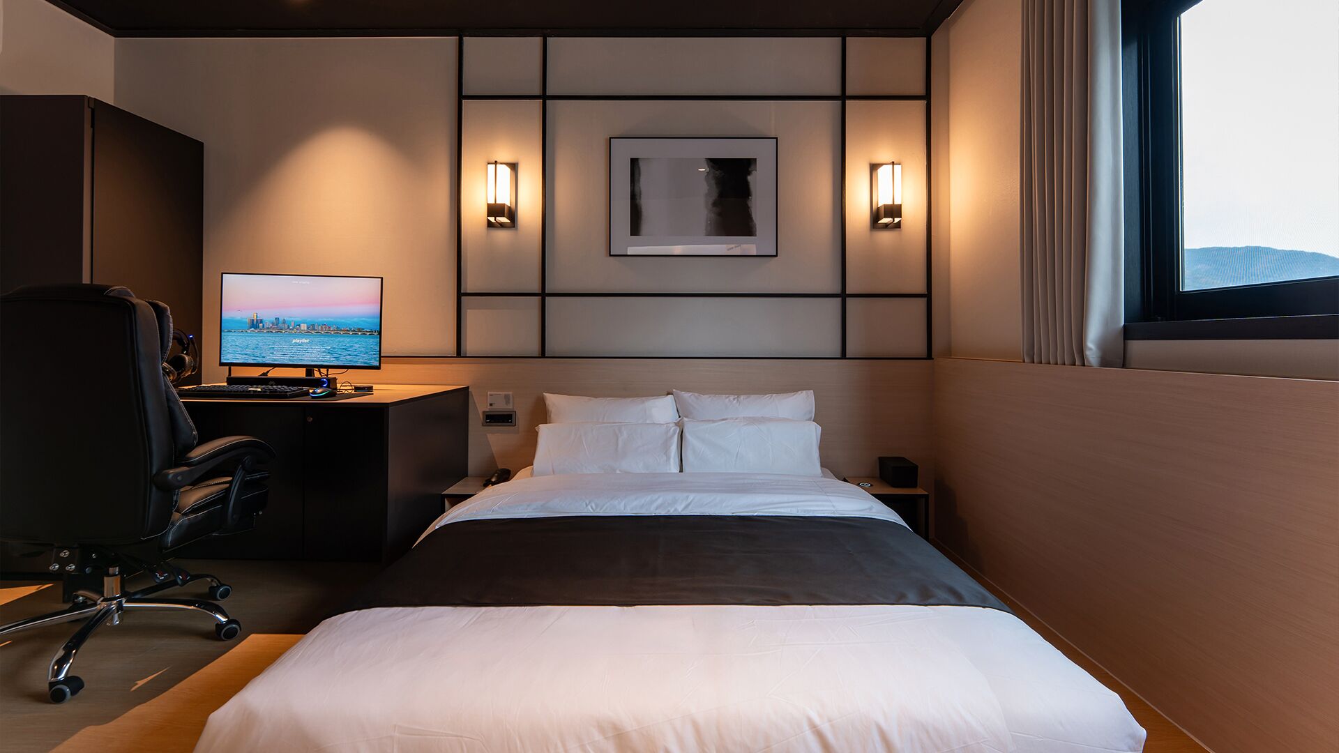 A hotel room with a bed, a desk with a computer, a chair, and a view of the city through the window.