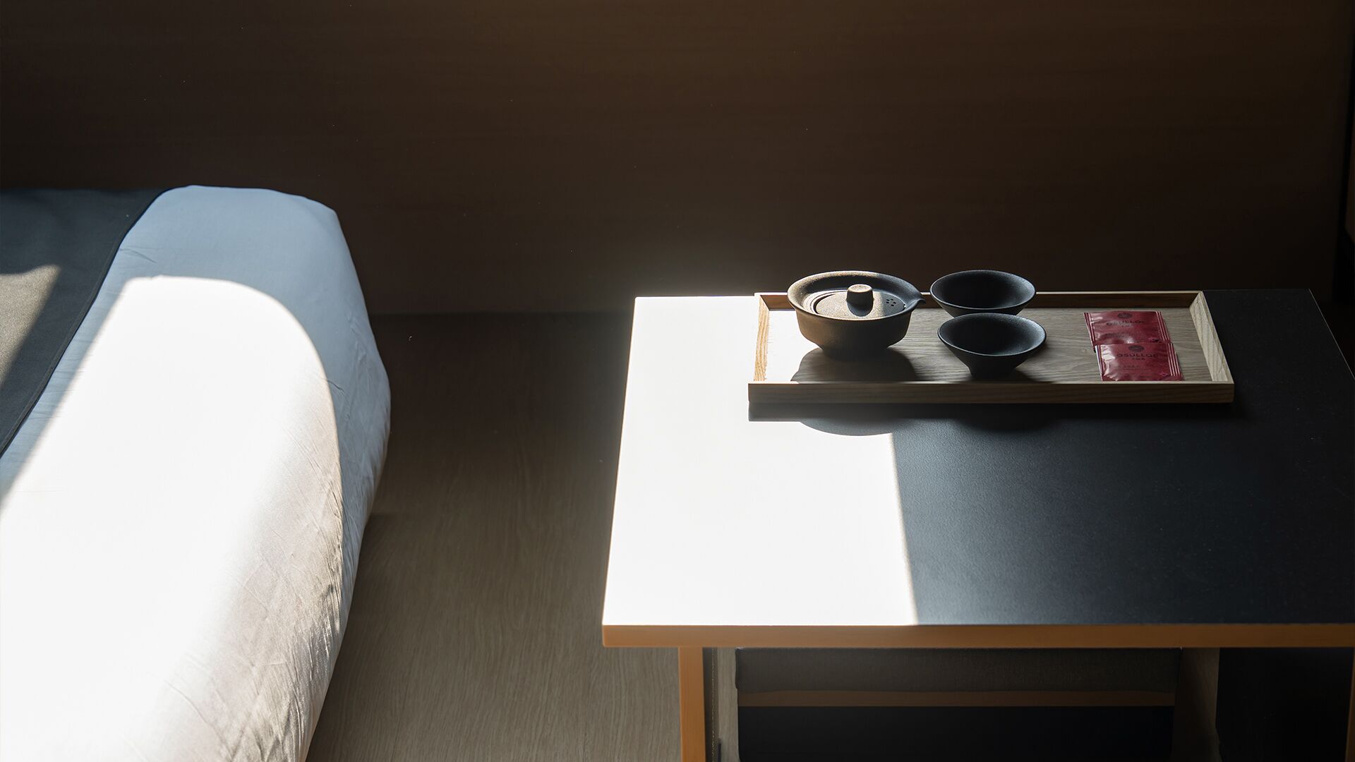 A neatly made bed with a blue cover, a small table with a tray holding a teapot, two bowls, and a packet of tea.