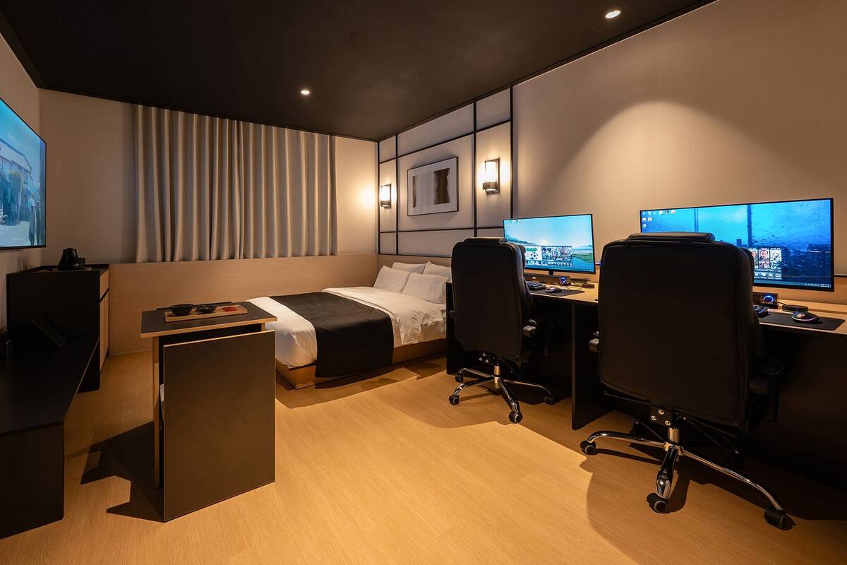 Ett modernt hotellrum med en säng, ett skrivbord, två datorskärmar och en tv.