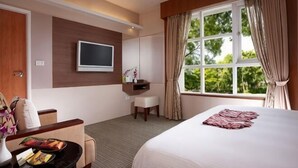 A hotel room with a bed, a desk with a chair, a TV mounted on the wall, a window with curtains, and a view of greenery outside. - Hwa Ger Hot Spring Hotel (Yilan)