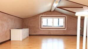 Une pièce avec un plafond en pente, une fenêtre à cadre blanc, une commode blanche et un ventilateur.