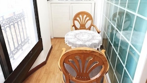 Une pièce d’angle avec une chaise en bois, une petite table ronde recouverte d’une nappe en dentelle, une armoire blanche et une fenêtre donnant sur un immeuble.