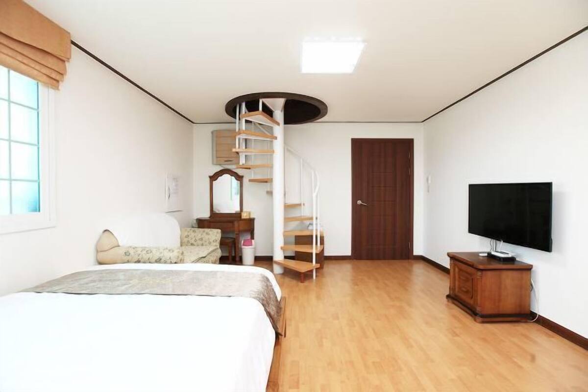 A bedroom with a bed, a TV, a wooden dresser, a desk with a chair, a mirror, and a spiral staircase.