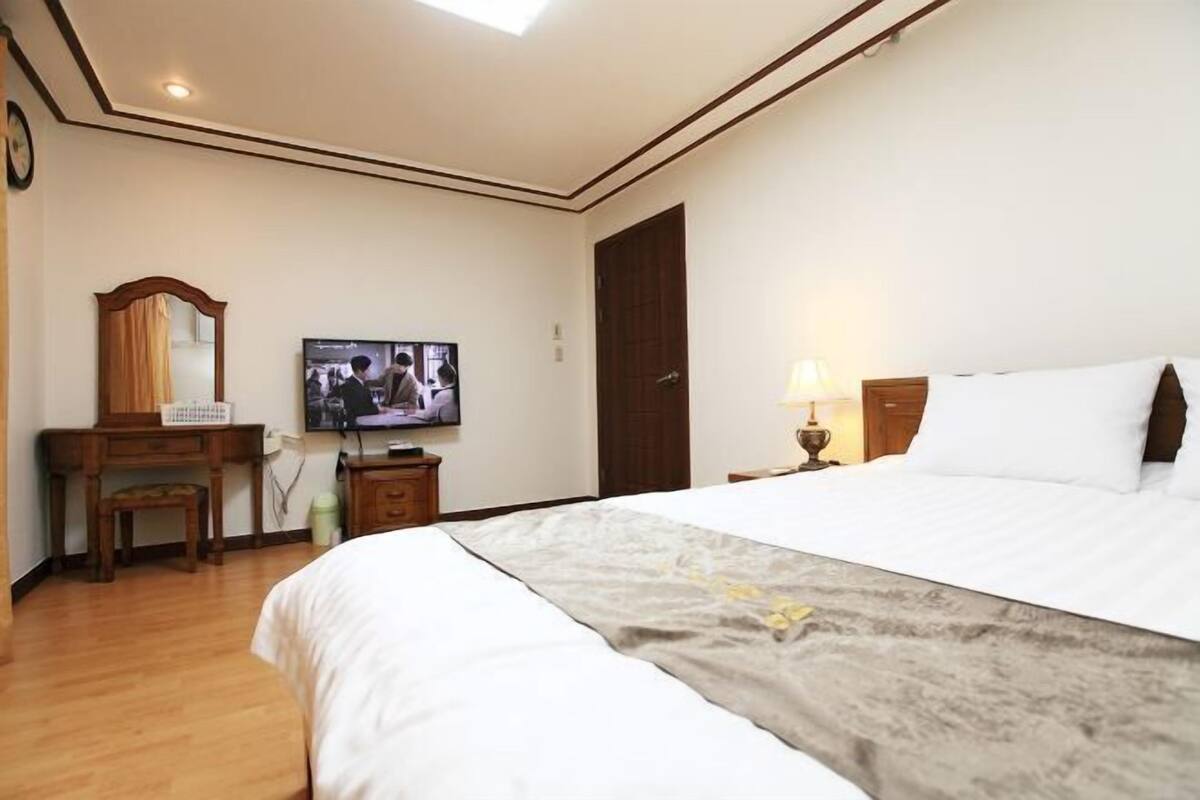 A hotel room with a large bed, a television on the wall, a desk with a chair, and a wooden wardrobe.