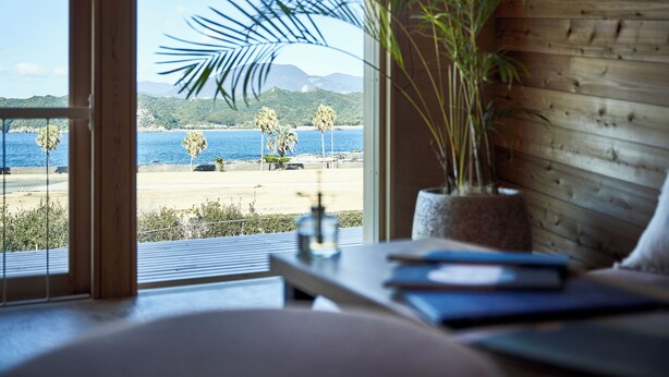A room with a large window overlooking a beach and mountains. There is a wooden wall, a plant, and a table with books and a bottle. - glampoceanluxe umikumano (Taiji)