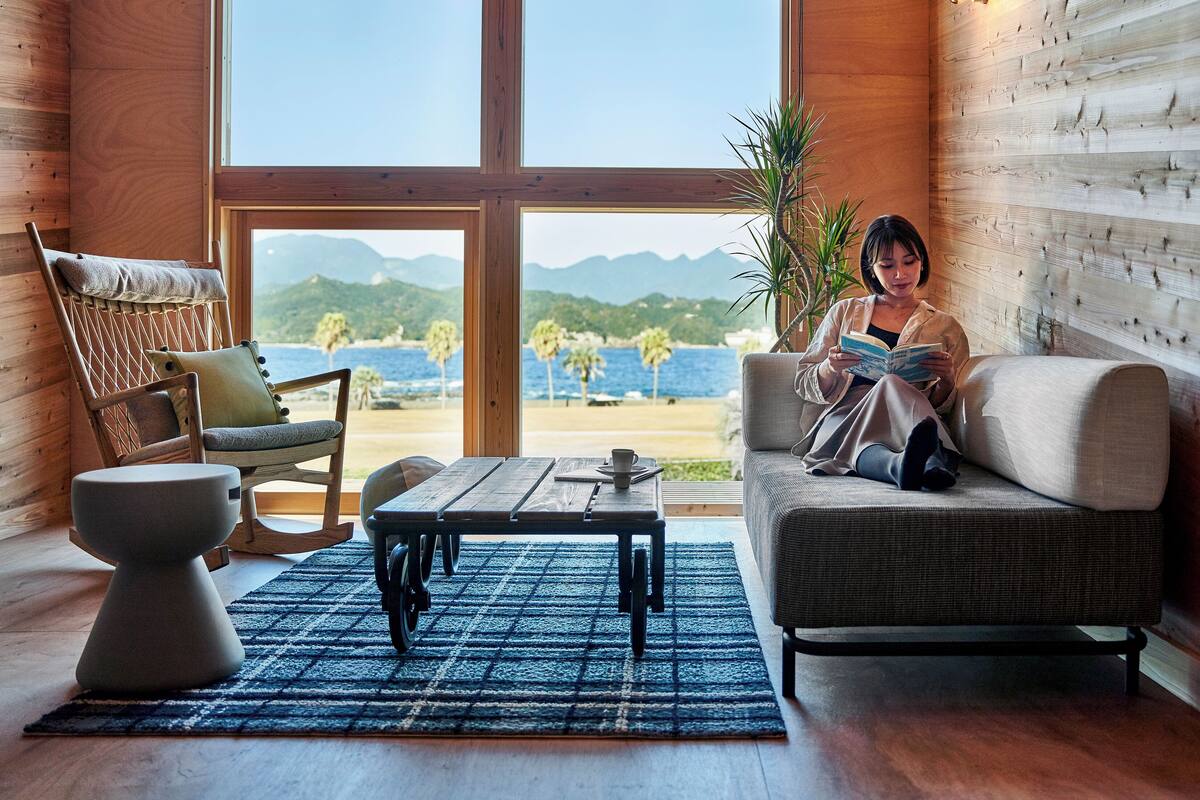 Una habitación acogedora con un ventanal grande, un sofá de mimbre, una mesa de madera y vistas a la playa y las montañas.