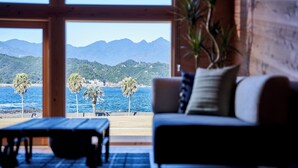 A room with a wooden interior, a sofa with cushions, a small table, and a view of the sea and mountains through large windows. - glampoceanluxe umikumano (Taiji)