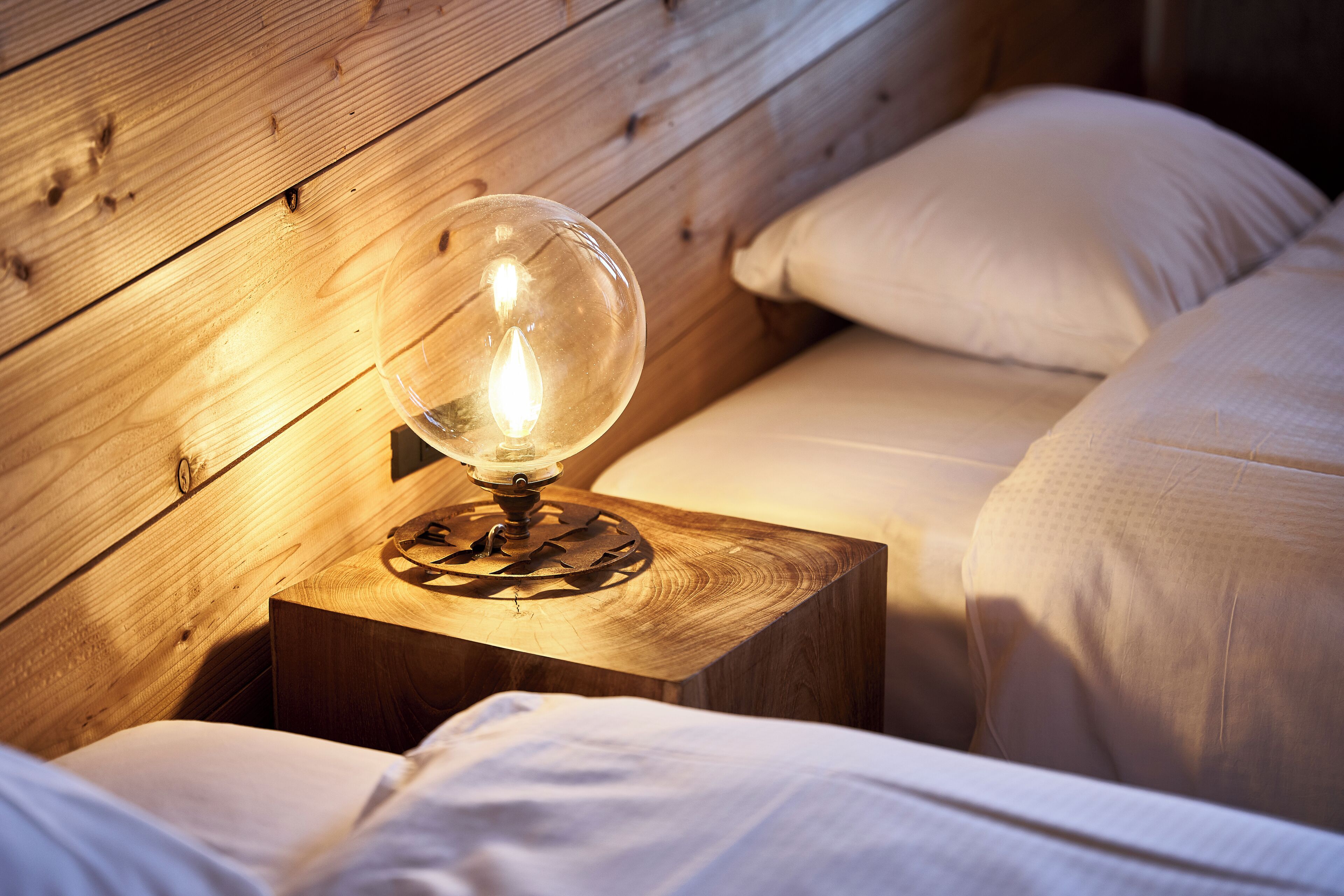 A wooden bedside table with a clear glass lamp, a wooden headboard, and white bedding.