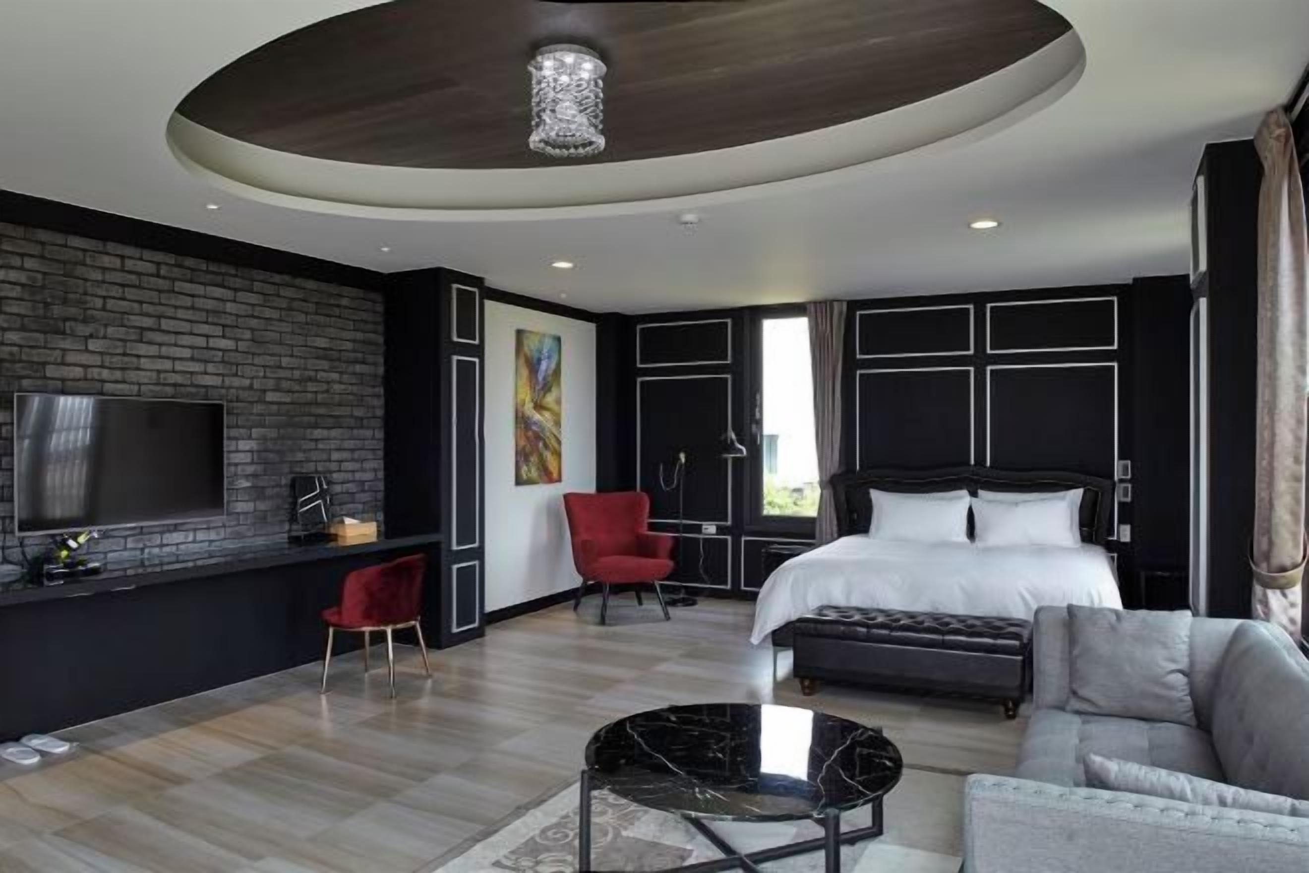 A modern hotel room with a large bed, a TV, a red chair, a sofa, and a unique ceiling design.