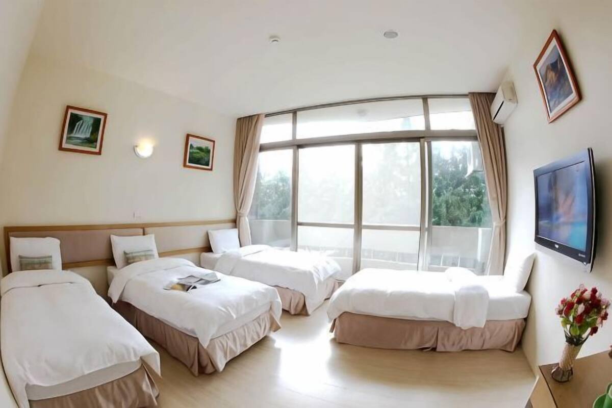 A hotel room with three beds, large windows, a flat-screen TV, and a vase with flowers.