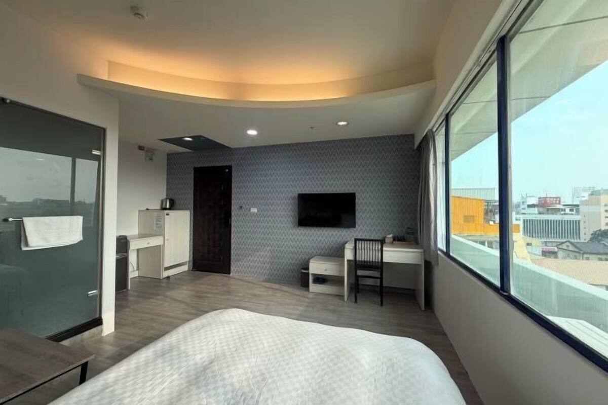 A modern hotel room with a bed, a desk, a chair, a TV, and a large window offering a city view.