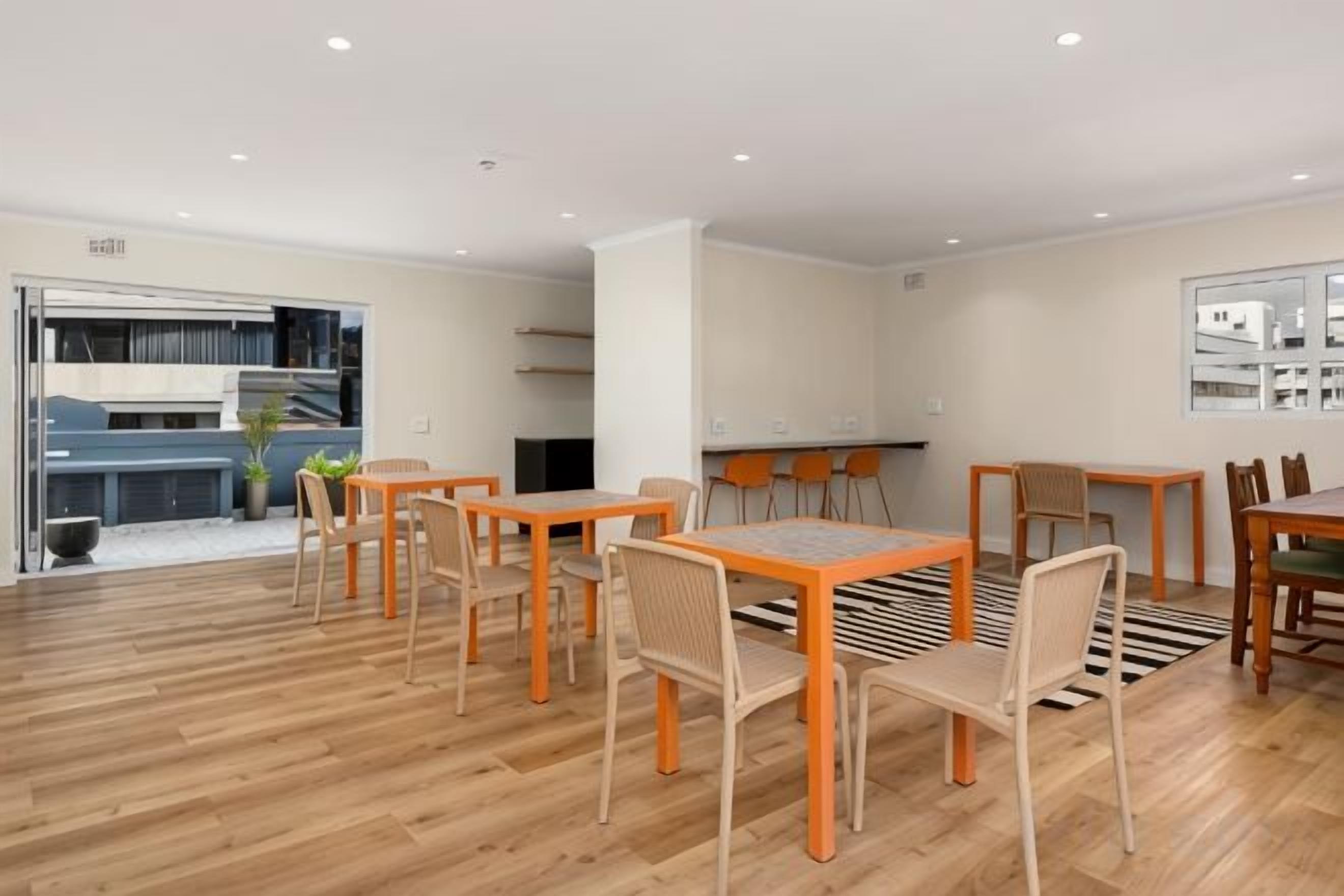 A spacious dining area with wooden flooring, multiple tables and chairs, and a view of a kitchen and balcony.