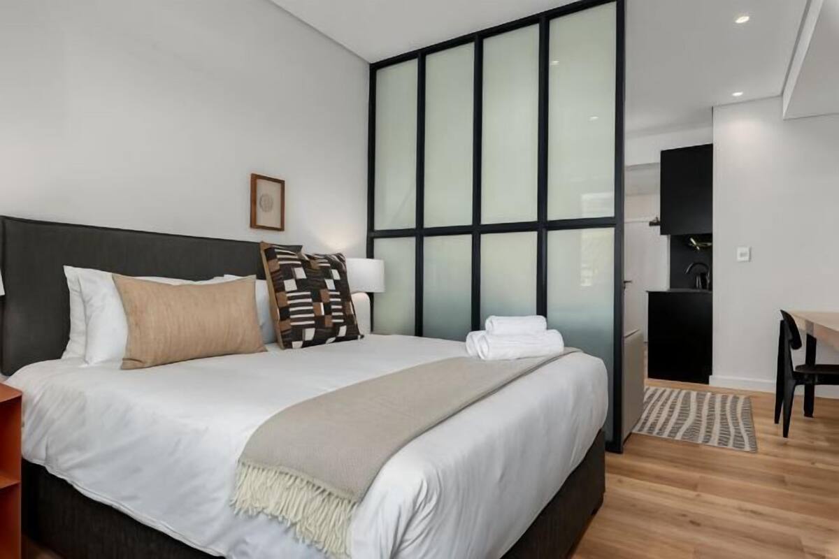 Ein modernes Hotelzimmer mit einem großen Bett, einem Schreibtisch, einem Sessel und Blick durch ein großes Fenster.
