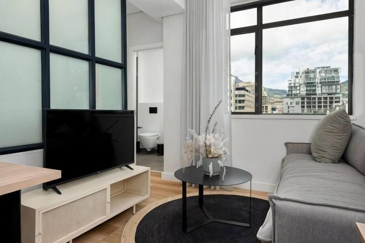 Ein modernes Wohnzimmer mit Flachbildfernseher, einer Couch, einem Couchtisch und Blick auf Gebäude durch große Fenster.