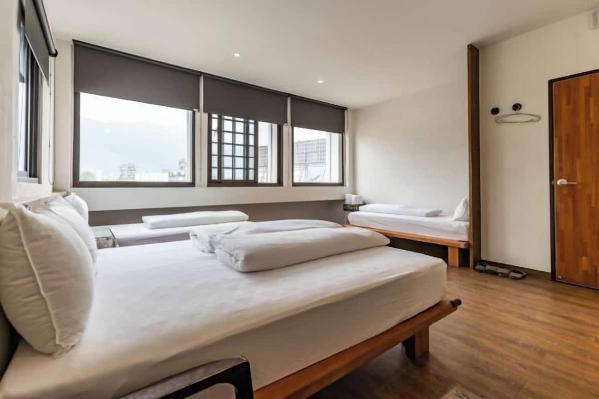 A modern hotel room with a large bed, a smaller bed, a wooden floor, and large windows with blinds.