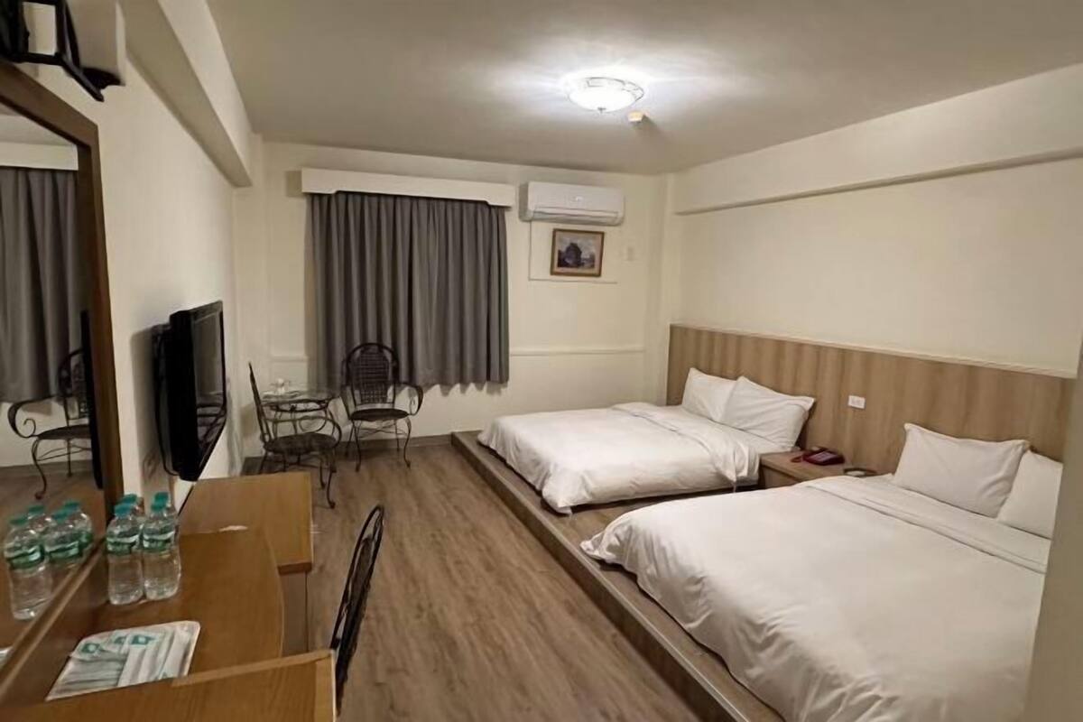 A hotel room with two beds, a desk with a chair, a TV, and an air conditioning unit.