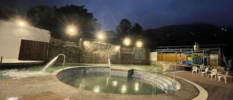 A heated outdoor pool area with a wooden deck, surrounded by stone walls and illuminated by overhead lights.