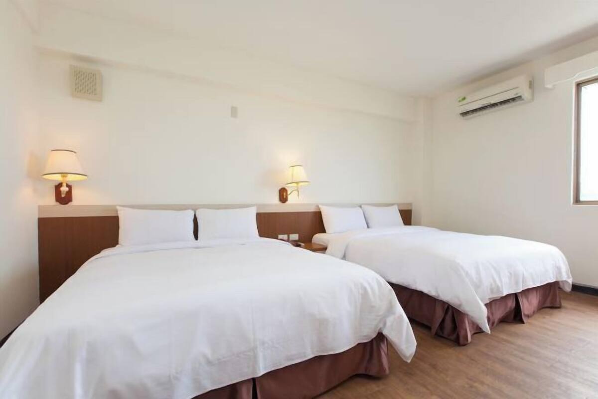 A hotel room with two beds, each with a headboard, bedside tables, lamps, and an air conditioning unit.
