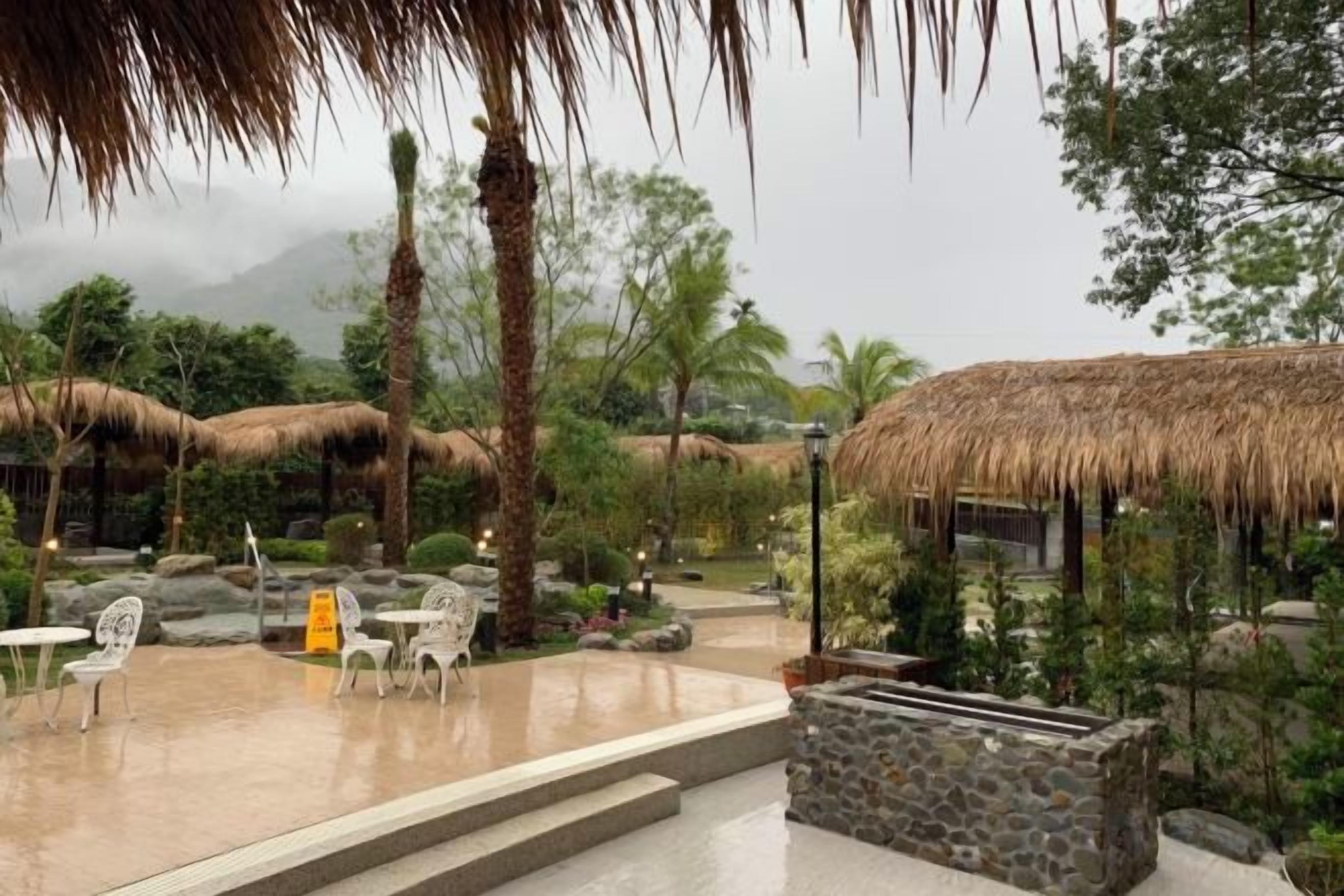 Un área de estar al aire libre con sombrillas de paja, muros de piedra y plantas tropicales.