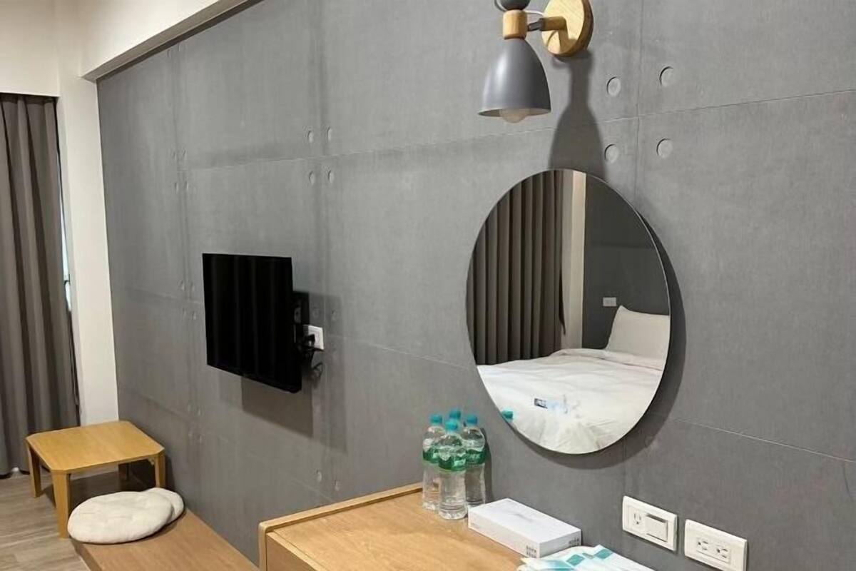 A modern hotel room with a grey wall, a round mirror, a flat-screen TV, a wooden bedside table, a small stool, and a bedside lamp.