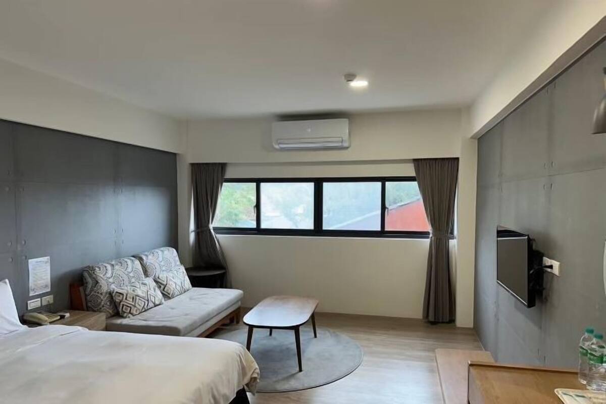 A modern hotel room with a sofa, two beds, a small table, a TV, and a window with curtains.