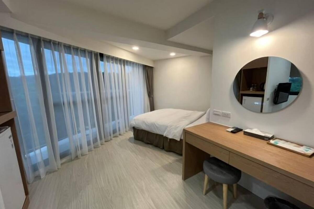 A hotel room with a bed, a desk, a chair, a round mirror, and a window with curtains.