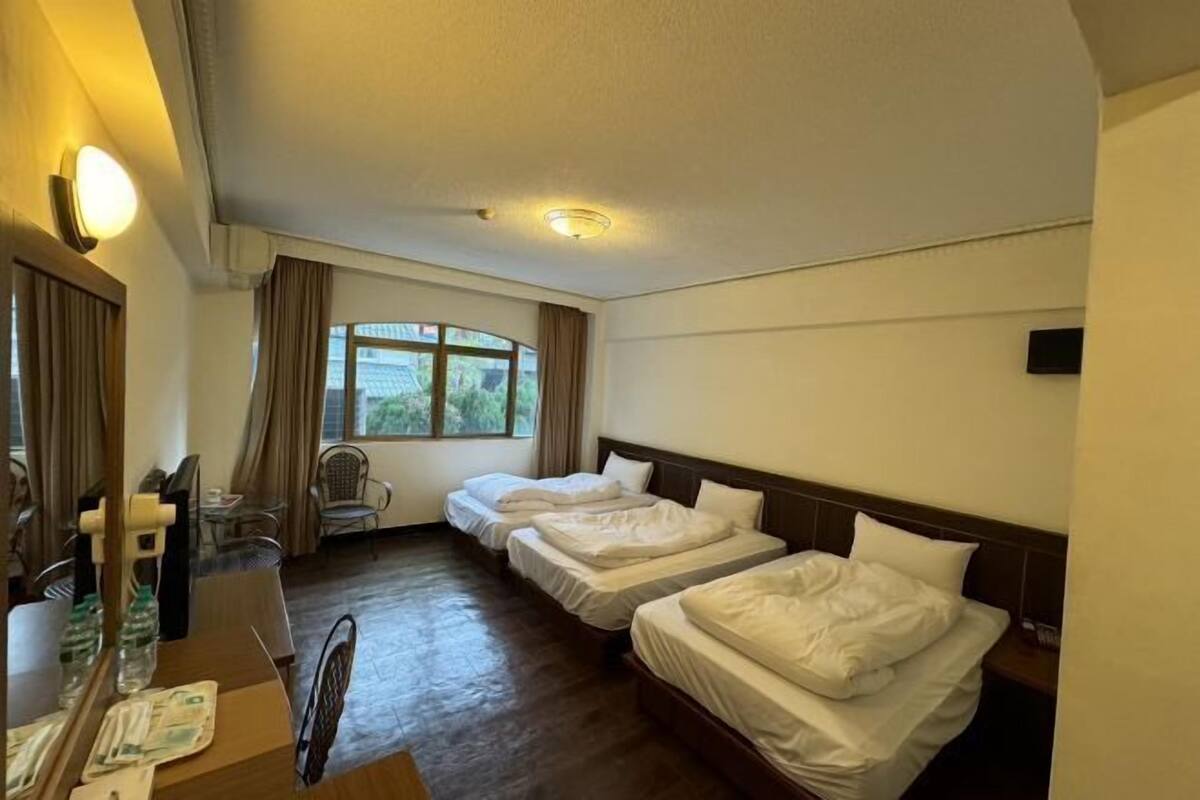 A hotel room with two single beds, a desk with a chair, a small table with a chair, a window with curtains, and a wall-mounted TV.