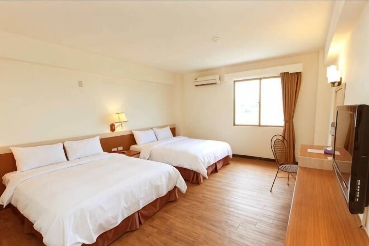 A hotel room with two beds, a TV, a desk, and a window with curtains.