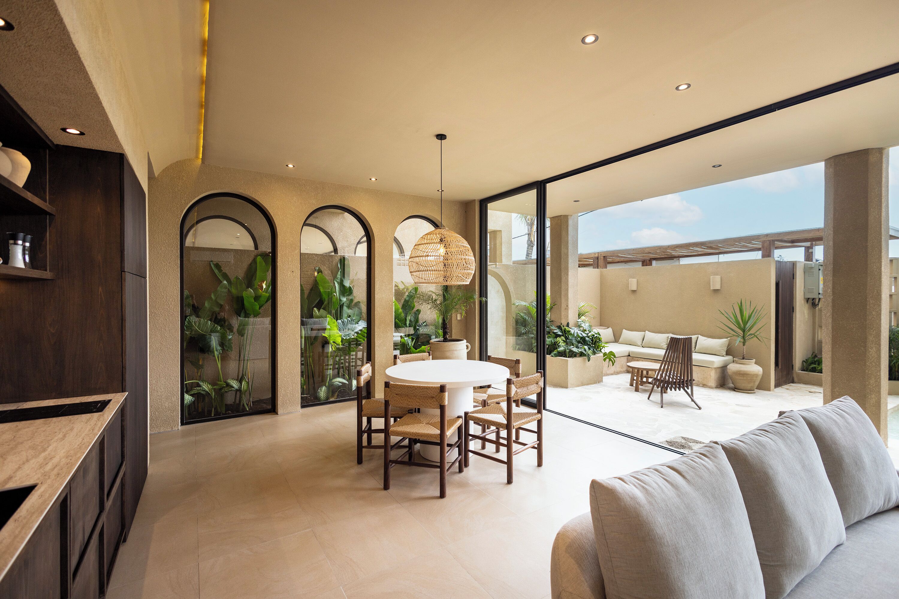 A modern interior with a round dining table, wicker chairs, a sofa, and large windows offering a view of the outdoors.