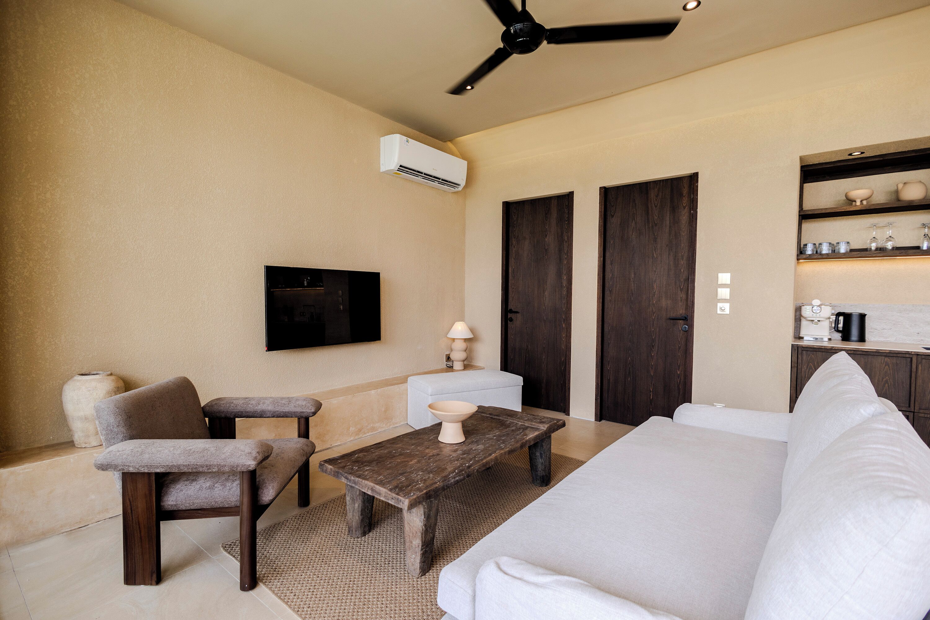Una habitación de hotel moderna con un sofá, dos sillones, una mesa de centro de madera, un televisor de pantalla plana, un ventilador de techo y un mini bar.